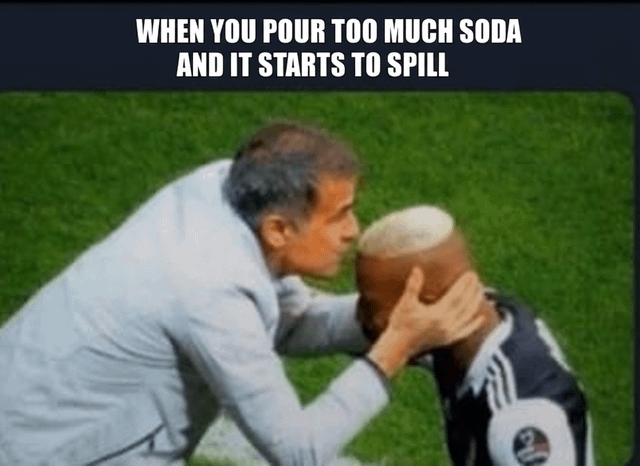A meme featuring a photograph of two individuals on a grassy sports field. The top text reads 'WHEN YOU POUR TOO MUCH SODA AND IT STARTS TO SPILL'. One person, wearing a light gray jacket, leans forward and firmly grips the head of another individual in a dark sports jersey with white stripes. The humor lies in the exaggerated, urgent grip being compared to the frantic attempt to manage an overflowing soda.