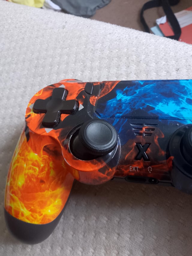 This is a humorous gaming meme centered on a custom third-party PlayStation-style controller with a red and orange flame print on the left side, and blue flame print on the right side. The left analog stick of the controller has completely sunken into the controller housing, leaving only the textured top cap visible sitting flush against the controller body. The joke exaggerates the common 'stick drift' hardware issue (where worn controller analog sticks register unwanted input without being touched) to an absurd extreme, implying the drift is so severe that the stick itself has collapsed into the device. The controller rests on a light gray textured fabric surface, likely a couch or armrest.