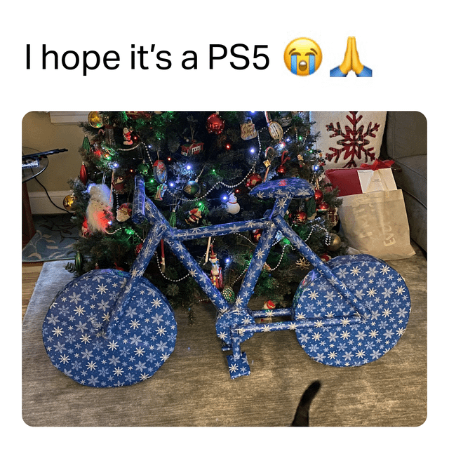 The image is a holiday-themed meme featuring a humorous contrast between gift expectations and reality. At the top of the image, there is text reading 'I hope it's a PS5 😭🙏' accompanied by a crying face emoji and praying hands emoji, expressing eager anticipation for a PS5 gaming console. Below this text is a photograph of a decorated Christmas tree adorned with colorful ornaments, lights, and a snowflake decoration in the background. In front of the tree sits a bicycle wrapped in blue wrapping paper with white snowflake patterns, clearly intended as a holiday gift. The meme plays on the common scenario of hoping for an exciting, high-demand gift (like a PS5) but receiving a more practical or underwhelming item (the bicycle), evoking a mix of humor and lighthearted disappointment.