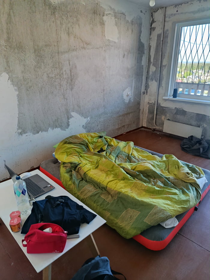 The image displays the interior of a partially gutted Soviet-era 3-room apartment that has not received renovations since 1983. All old wallpaper has been stripped, leaving rough, unfinished concrete walls. The resident, who recently went through a breakup, is starting their new life here with minimal belongings: an inflatable air mattress with a crumpled green comforter serves as a temporary bed, a small white folding table holds a laptop, bottle of water, clothing items, a red toiletry bag, and small containers. A window with metal security bars looks out over a distant residential area, with a radiator positioned underneath it, and a bare lightbulb hangs from the ceiling. The poster notes they have already completed 3 days of work removing the old wallpaper, and are documenting their step-by-step renovation process while asking for well wishes.