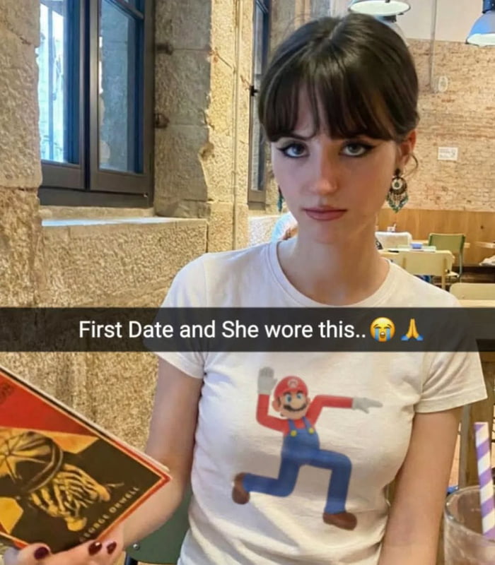 The image depicts a woman seated in a restaurant during a first date. She is wearing a white t-shirt featuring Mario, the iconic character from Nintendo's Super Mario franchise, in a running pose with arms outstretched. The woman holds a menu in her left hand, and the background shows a casual restaurant setting with stone walls and wooden furniture. Overlaid text at the top reads 'First Date and She wore this..😂🙏', combining a laughing emoji and a folded hands emoji, suggesting the poster finds humor or relief in the shared interest indicated by the Mario shirt.