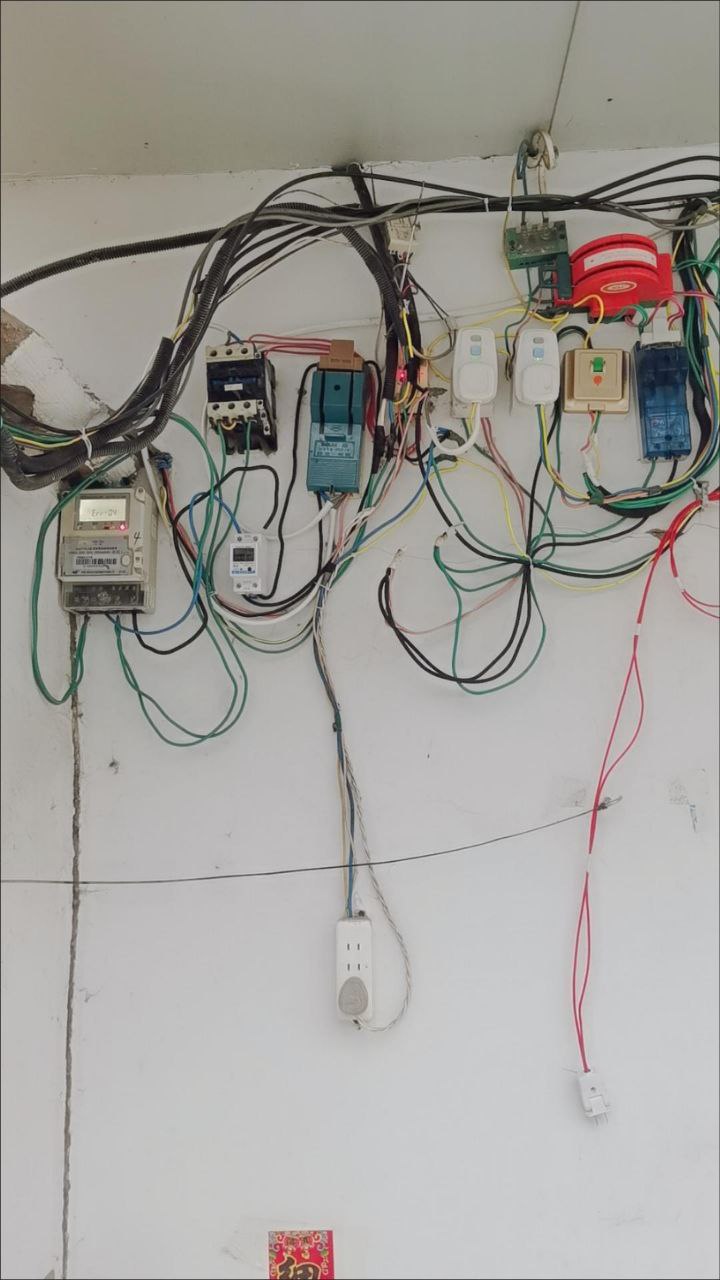A photograph showing an extremely disorganized and potentially dangerous electrical setup on a white wall. Multiple electrical components including circuit breakers, meters, switches, and outlets are visible with numerous multicolored wires (red, green, yellow, blue, black, white) haphazardly connected and hanging loosely. The wiring appears to lack proper organization or safety standards. A small red and gold decorative item with Chinese characters is visible at the bottom, suggesting this might be in a Chinese-speaking region.