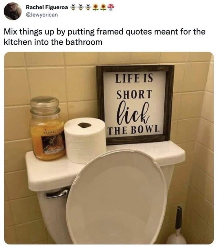 This is a screenshot of a post from X (formerly Twitter) that shares a humorous home decor prank. The post text suggests making home decor more interesting by placing kitchen-themed framed quotes in the bathroom. The accompanying photo shows a bathroom toilet with a roll of toilet paper and a Yankee Candle sitting on the toilet tank, next to a framed sign. The sign reads 'LIFE IS SHORT lick THE BOWL', which is a common wholesome kitchen quote referring to licking leftover food batter from mixing bowls. When placed in the bathroom next to a toilet, the quote takes on a crude, awkward double meaning referring to licking the toilet bowl, creating the core comedic effect of the meme.