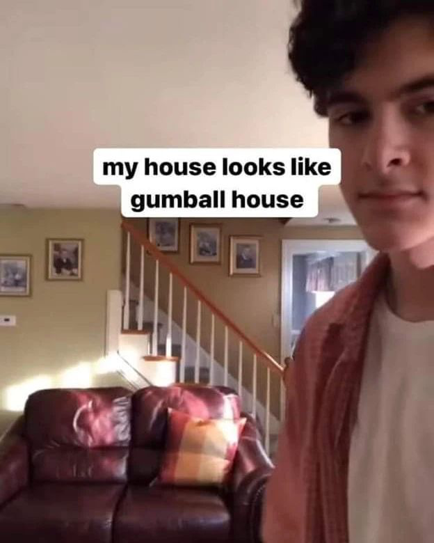 A partially visible young man stands on the right edge of the frame taking a selfie showcasing his home's living room. The background features a dark burgundy leather couch with a plaid throw pillow, a staircase with white balusters and a light wood handrail, pale green walls decorated with multiple framed portrait photographs, and a doorway to another room. White text with black font is overlaid on the upper center of the image, reading 'my house looks like gumball house', pointing out the striking similarity between this real living room layout and the interior of the Watterson family home from the popular animated series *The Amazing World of Gumball*, creating a humorous real-life parallel to the fictional cartoon setting.