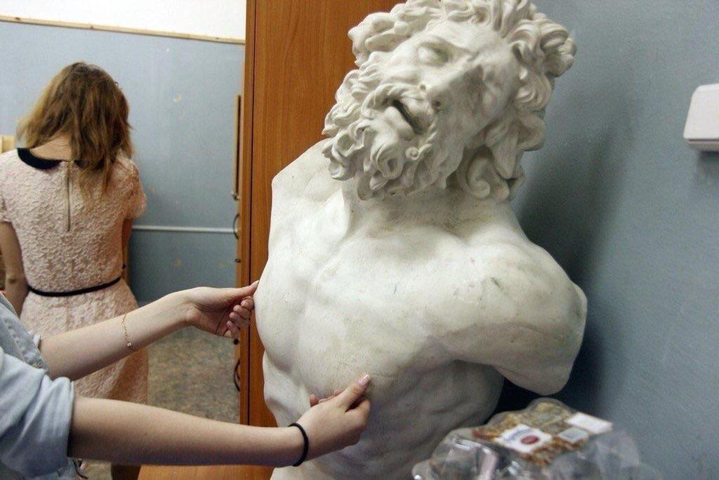 This photograph features a white marble bust of the classical Greco-Roman figure Laocoön, identifiable by his signature curly hair, full beard, and iconic anguished, tilted-head expression with his mouth open as if crying out. Two arms belonging to an off-frame person wearing a light blue shirt are extended to squeeze and prod the pectoral muscles of the statue. In the background, a woman with light brown hair in a pink lace dress stands facing away from the camera next to a wooden door and grey painted interior walls. A small packaged food product is visible on a surface at the bottom right of the frame. The image functions as a meme due to the humorous juxtaposition: the statue's existing over-the-top pained expression appears to be a direct, exaggerated reaction to the irreverent, inappropriate act of touching the classical artwork. There is no overlaid text on the image.