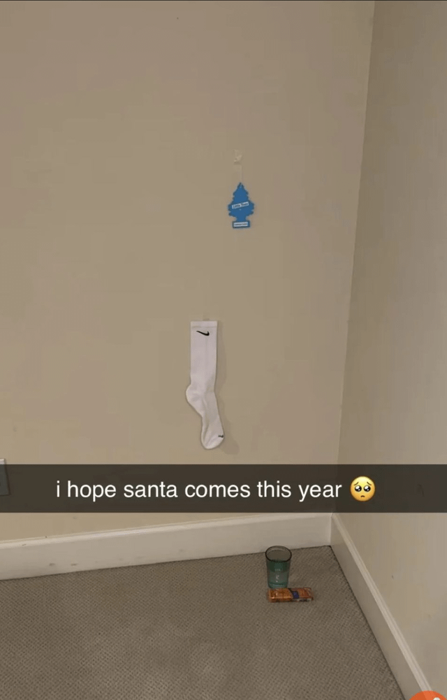 A photograph of a corner in a room with beige walls. On the wall, a blue fire hydrant-shaped decoration is hung with a string, and directly below it, a white Nike sock is also hung, resembling a makeshift Christmas stocking. On the carpeted floor in the corner, there is a clear glass cup and a small snack bar. A text overlay at the bottom reads 'i hope santa comes this year 😢' (with a sad emoji), creating a humorous and lighthearted scene where the sock is humorously positioned as a stocking, expressing hope that Santa will notice it despite its unconventional location.
