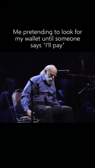 This meme consists of a white text caption on a solid black background at the top, followed by an image of an elderly bald man with a long white beard, wearing a blue collared shirt and dark pants, sitting on a chair on a dark stage and looking down towards his lap as if searching for something. The caption reads 'Me pretending to look for my wallet until someone says 'I'll pay'', using the man's searching posture to humorously visualize the common relatable behavior of pretending to fumble for one's wallet when a group bill arrives, waiting for another person to volunteer to cover the cost.