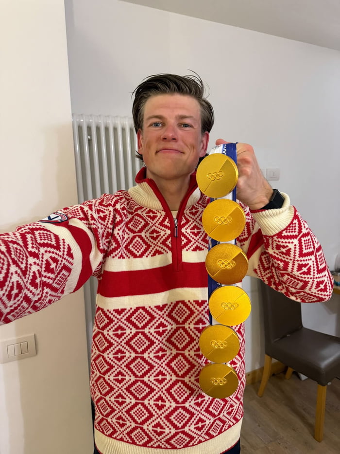 The image features Johannes Thingnes Bø, a highly decorated Norwegian biathlete, taking a playful selfie. He uses a forced perspective/reflective trick with mirrors to create the illusion of holding six Olympic gold medals strung on a single blue ribbon; in reality, it is one medal reflected multiple times. He wears a traditional red-and-white Norwegian patterned knit sweater, has a proud, subtly amused facial expression, and holds the top of the medal ribbon up to the reflective surface to achieve the visual gag. The background shows an indoor residential space, including a white radiator, wall light switch, and a dark upholstered chair with wooden legs.