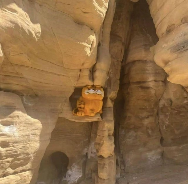 This photograph shows an orange Garfield plush toy sitting wedged into a small ledge crevice between large tan sandstone rock formations in a canyon. The plush has Garfield's iconic half-lidded bored expression, and its orange color blends in closely with the surrounding rock. The plush appears to have been lost or abandoned here, trapped out of reach in the rock formation.