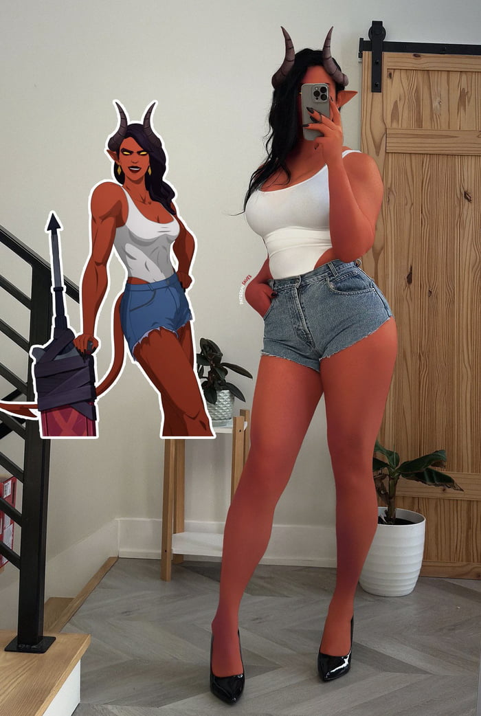 The image features a side-by-side comparison between an animated demon character and a real person cosplaying as the character. The animated character has red skin, long black hair, prominent horns, a white tank top, blue denim shorts, and is holding a wrapped weapon with a red 'X' on it. She stands confidently with one hand on her hip. To her right, the real person mirrors the character's outfit: a white tank top, blue denim shorts, and has red skin (likely body paint) with horns. She is taking a selfie in a room with a wooden door, a small plant stand, and a staircase visible in the background.