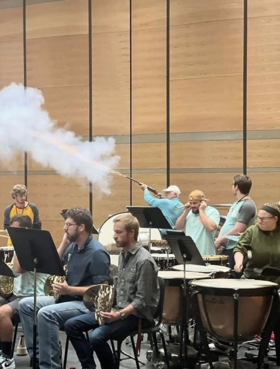 The image captures an absurd scene at an indoor concert band or orchestra practice session, held in a room with light wood panel walls. Several seated musicians hold instruments including French horns and sit in front of timpani drums, most displaying bored, unphased expressions as they look at their sheet music stands. In the background, a man wearing a light blue shirt and white baseball cap stands and fires a flare gun, which emits a large trail of white smoke. One man standing near the flare gun user covers his ears in anticipation of loud noise, while the rest of the group shows little to no reaction to the extremely out-of-place, chaotic action. The humor derives from the stark contrast between the mundane, calm routine of music practice and the absurd disruptive event, paired with the understated reaction of most attendees.