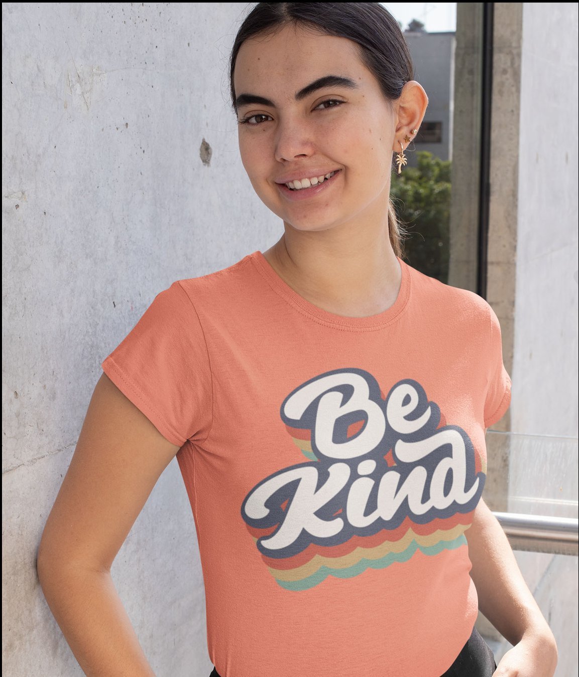 The image shows a young woman with dark hair pulled back in a ponytail, smiling warmly at the camera while leaning against a light grey concrete wall outdoors. She is wearing a coral peach short-sleeved t-shirt featuring a retro-style graphic that reads 'Be Kind' in white cursive lettering with a dark navy outline, accompanied by layered wavy stripes in orange, yellow, and teal under the text. This t-shirt design is part of a charity merchandise line created to raise funds to support people in distress.