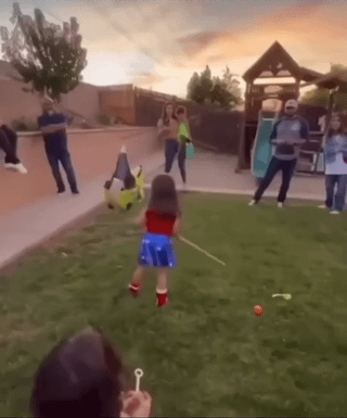 The image captures an outdoor park scene at sunset, using a forced perspective optical illusion to create a funny visual gag. A person in the immediate foreground holds a small plastic bubble wand pointed directly at a young girl in a red top and blue sparkly skirt standing further away on a grassy lawn. The exact alignment of the wand and the girl makes it appear as if the girl is tiny and trapped inside a giant bubble being blown from the wand. Other casual park visitors are visible in the background, including one person who has fallen over on the sidewalk, contributing to the lighthearted, casual atmosphere. The accompanying title frames the illusion as a silly, 'sweet' method to get candy, playing on the idea of catching a child as if they were a treat inside a bubble.