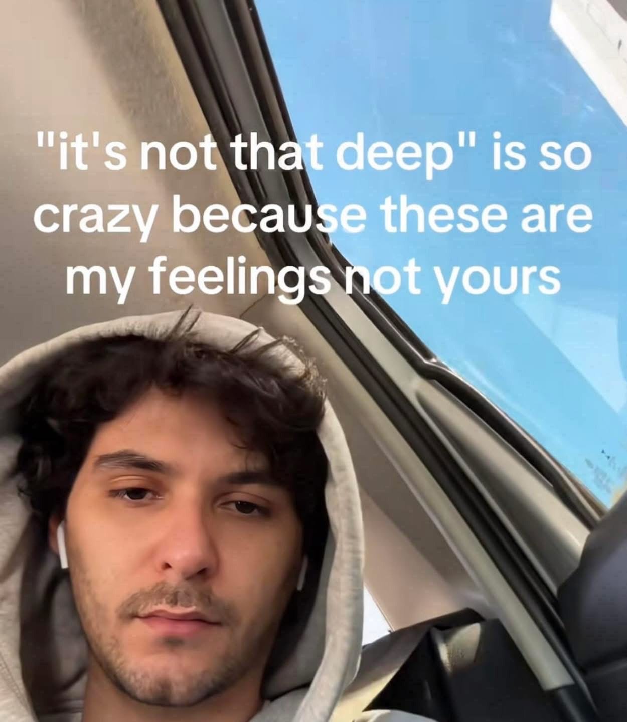 This is a single static reaction meme featuring a real young man with dark curly hair, light facial stubble, wearing a gray hoodie with the hood pulled up and white wireless earbuds, sitting inside a car. A clear bright blue sky is visible through the car window beside him. He has a deadpan, unamused, tired expression as he looks directly at the camera. White overlaid text on the upper portion of the image reads: "'it's not that deep' is so crazy because these are my feelings not yours". The meme conveys frustration at the common experience of other people minimizing or invalidating one's personal emotional experiences.