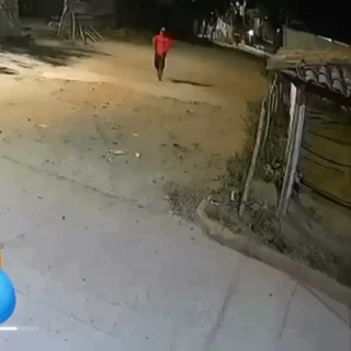 This is a low-resolution security camera screenshot captured at night, showing a lone person wearing a bright red top and dark pants walking away down a dimly lit, empty unpaved rural or suburban street. Simple makeshift shacks line the right side of the road, and no other people or vehicles are present in the scene. The accompanying title 'bro doesn't fear' frames the content as a humorous meme about the person's apparent lack of fear of being alone in an isolated, dark, potentially unsafe area after dark.