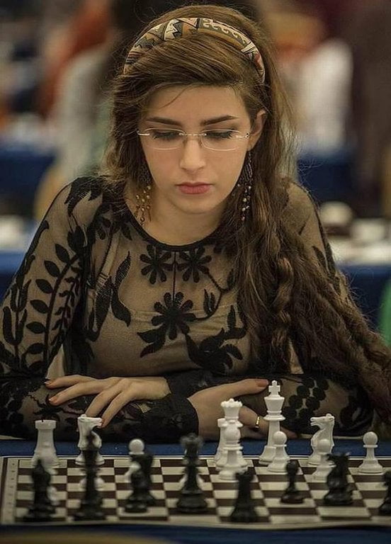 The image depicts Dorsa Derakhshani, a chess player, seated at a chessboard in a tournament setting. She has long brown hair adorned with a patterned headband, wears glasses, and a sheer black top with floral embroidery. Her hands rest near the chess pieces, and she appears focused on the game. The background is blurred, emphasizing the competitive environment. The context highlights her defiance of the Islamic regime's hijab ultimatum, subsequent success as a U.S. team member, Chess Grandmaster, world champion, and medical school enrollment.