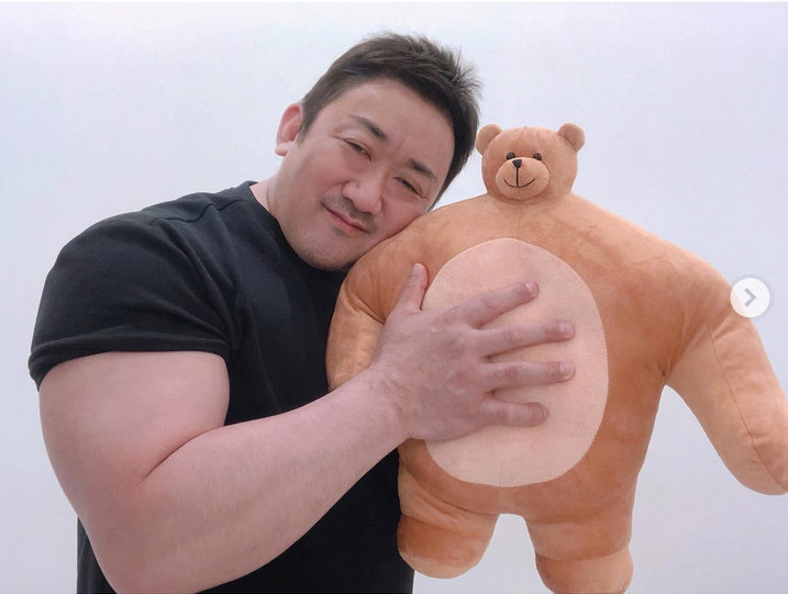 A muscular individual wearing a black short-sleeved shirt is seen hugging a large, chubby brown teddy bear with a light beige circular belly. The person has well-defined arm muscles and is gently pressing their hand against the teddy bear's belly, creating a charming contrast between their physical strength and the soft, cuddly nature of the toy. The background is a plain white, keeping the focus entirely on the warm interaction. A small circular arrow icon on the right edge suggests this image might be part of a carousel or slideshow, a common feature in social media posts.