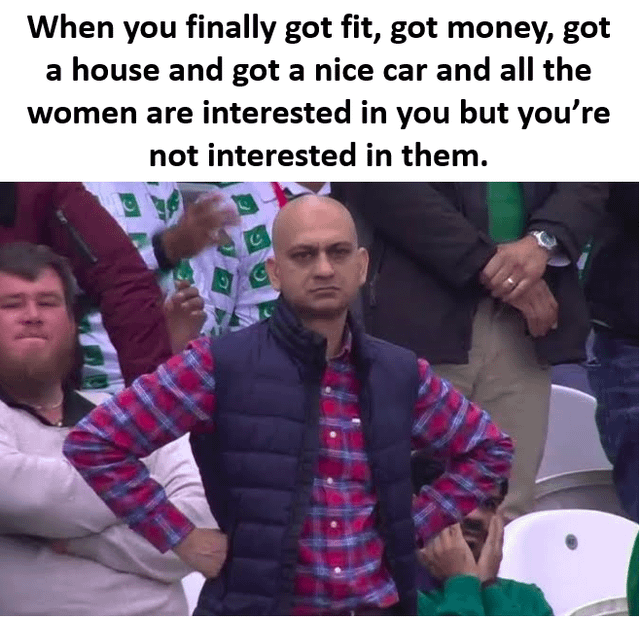 This meme is made of text placed above a well-known viral reaction photo. The top text outlines a relatable scenario: a person has reached major common life goals, including getting fit, becoming financially stable, owning a house, and owning a nice car, which makes them desirable to women, but they have no romantic interest in women at this point in their life. The image below the text features a bald South Asian man standing in a stadium crowd, wearing a dark puffer vest over a red and blue plaid shirt, with both hands resting on his hips. He has a completely unimpressed, apathetic, unbothered facial expression that perfectly aligns with the uninterested sentiment described in the text.