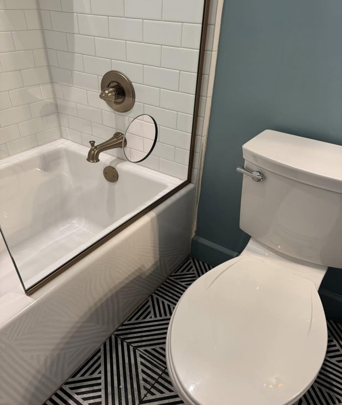 A photograph of a modern bathroom featuring a white bathtub with a glass shower door, which has a prominent circular hole near the faucet area. To the right of the bathtub is a white toilet with a silver flush handle. The floor has a black and white geometric zigzag pattern, while the walls are tiled in white (around the bathtub) and painted light blue (near the toilet). Brass fixtures, including the bathtub faucet and shower knob, are visible. The focal point of the image is the circular hole in the glass shower door, prompting the question about its purpose.