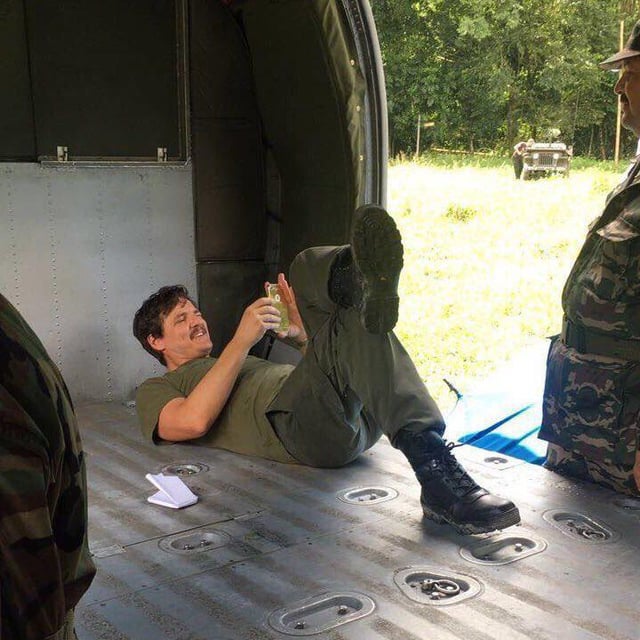 A meme image depicting a man in a green outfit lying casually on the metallic floor of a helicopter's open interior. He is holding a small, possibly glass bottle in his right hand, with his left hand supporting it, and his legs propped up, feet resting on the helicopter's side. His expression is amused and satisfied, with a slight smile. Surrounding him are individuals in military-style camouflage clothing: one stands to his right near the helicopter's entrance, and another is visible in the background near a vehicle. The setting is an outdoor grassy field with trees, suggesting a military or expedition context. The meme humorously uses the man's relaxed, self-assured posture to represent the relatable experience of re-reading one's own social media comment after receiving upvote notifications, embodying satisfaction from positive feedback.