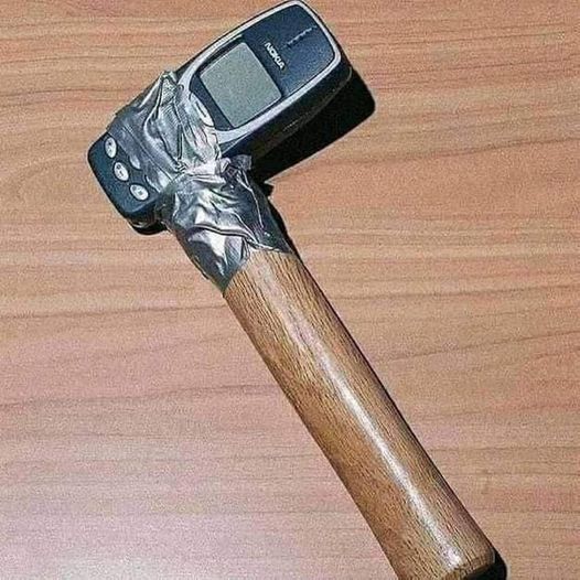 The image displays a humorous modified object: an old, widely considered indestructible model of Nokia candybar mobile phone is affixed as the head of a hammer to a polished wooden handle, secured with crumpled silver duct tape. The hybrid object rests on a light brown wood grain tabletop. The joke relies on the long-circulated internet meme that vintage Nokia feature phones are extremely durable, to the point that they could function as a hammer or withstand the same level of heavy impact as construction tools.