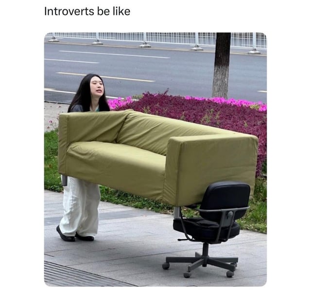 The meme opens with a top text caption reading 'Introverts be like'. Below the caption is a photograph of a young woman walking on a paved sidewalk outdoors, carrying a large, heavy full-size olive-green fabric couch completely by herself, with a visibly strained expression from the physical effort. A black rolling office chair is placed on the ground next to the couch, and the background shows a public road, purple and pink roadside flower beds, a tree, and road guardrails. The visual joke highlights the extremely relatable trait of introverts, and people who dislike asking others for favors, choosing to go to impractical, extreme lengths to complete difficult tasks entirely alone rather than requesting support from other people.