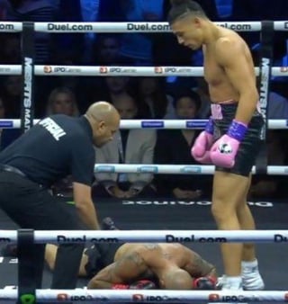 A photograph capturing a dramatic moment in a boxing match. Inside the ring, a referee bends down to check on a boxer who has fallen face-first onto the canvas. Another boxer, wearing pink gloves and trunks, stands nearby observing the scene. The ring is surrounded by a banner displaying 'Duel.com' at the top, and an audience is visible in the background behind the ropes.