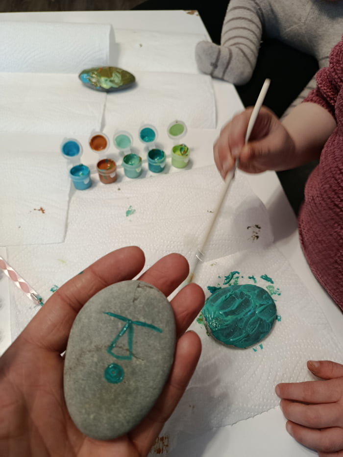 This image shows an adult and a child doing rock painting together at a table covered with protective paper towels. An adult hand holds up a smooth grey rock with a simple teal drawing that is intentionally shaped like a penis, which is the hidden joke of the post. In the background, small pots of acrylic paint are laid out, the child's hands are visible holding a paintbrush working on another rock that is already coated in teal paint. The original premise is that the parent made this hidden dirty drawing during the casual parent-child art activity, and his wife claimed no one would notice the inappropriate shape, while he was sure other internet users would spot it immediately.