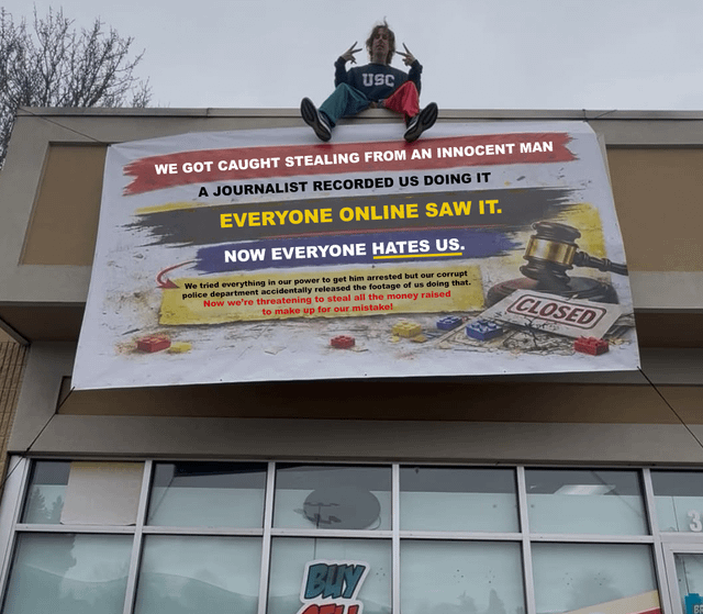 The image shows a large snarky protest banner hung over the storefront of a Bricks & Minifigs, a Lego resale chain location. The banner text accuses the store of stealing from an innocent man, being caught on tape by a journalist, the footage going viral leading to widespread public anger, then the store attempting to frame the victim only for police to accidentally release footage proving the store's wrongdoing, and now threatening to seize raised funds to cover their losses. The banner also includes graphics of Lego bricks, a judge's gavel, and a 'CLOSED' sign referencing the store's business and controversy. A young person wearing a USC sweatshirt and mismatched red and green pants sits on the roof edge above the banner, holding up double peace signs toward the camera.