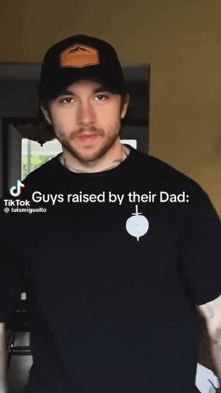 This image is a screenshot of a short TikTok video, showing a young adult man with light facial stubble wearing a black baseball cap with an orange mountain logo patch and a black t-shirt printed with a white sword and shield graphic. He stands facing the camera with a calm, neutral deadpan expression. Overlaid white text on the image reads "Guys raised by their Dad:", referencing a widely shared relatable stereotype that men primarily raised by their fathers often have a laid-back, low-drama, practical demeanor and casual, utilitarian personal style matching the vibe and appearance of the man in the clip.