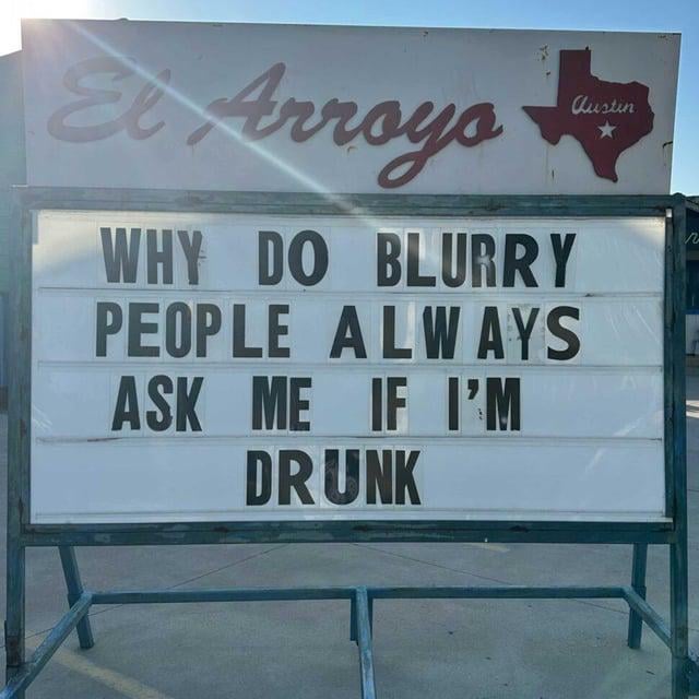 A photograph of an outdoor sign from 'El Arroyo' in Austin, Texas. The sign features a red Texas state outline with 'Austin' written inside at the top. Below, black text on a white background reads: 'WHY DO BLURRY PEOPLE ALWAYS ASK ME IF I’M DRUNK'. The sign is mounted on a metal frame, with sunlight creating a slight glare on the surface. The humor stems from the pun: 'blurry people' (those who appear blurry to the observer) asking if the observer is drunk, reversing the common scenario where drunk people see others as blurry.