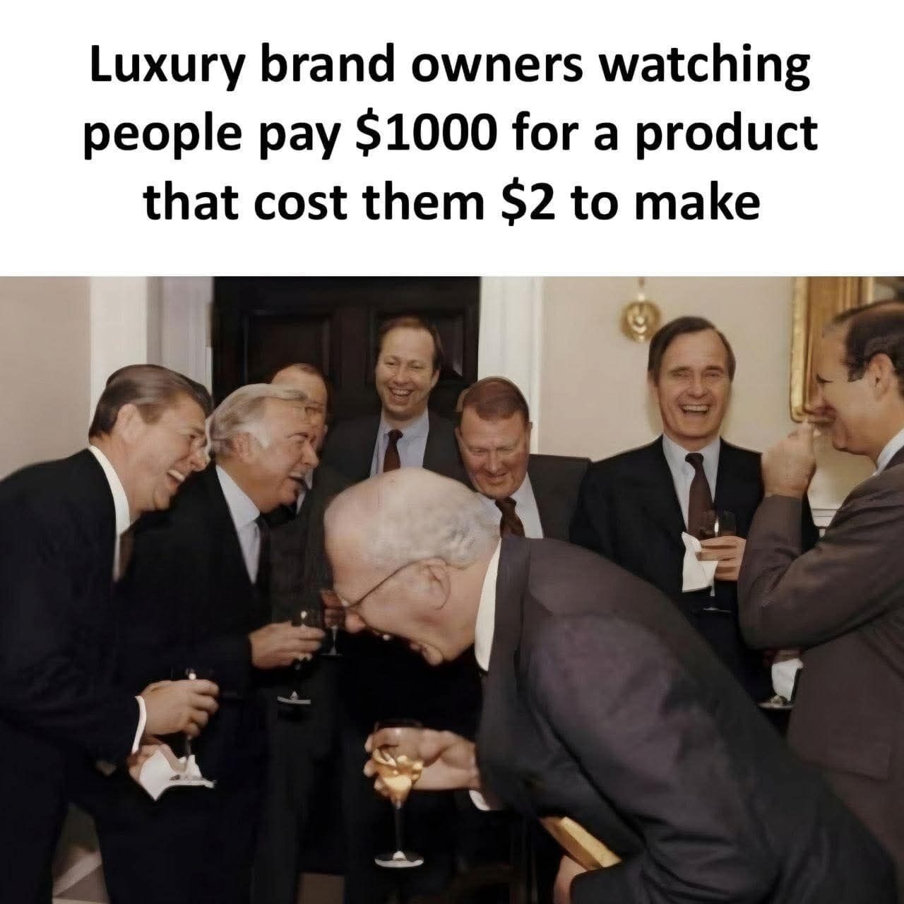 This is a single-panel satirical meme. The top of the image carries text that reads: "Luxury brand owners watching people pay $1000 for a product that cost them $2 to make". Below the text is a widely known historical photograph of a group of older men in formal suits at an upscale indoor event, holding wine glasses and laughing very heartily. One bald man in the foreground is bent over from how hard he is laughing, while the other men around him are also grinning or chuckling intensely. The joke critiques the extremely high markup of luxury goods, where consumers pay hundreds of times the actual production cost of products purely for the prestige of the brand name.