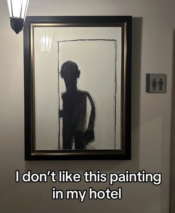 The image captures a framed black-and-white abstract painting mounted on a hotel hallway wall, adjacent to a standard public restroom sign indicating gendered facilities. The painting depicts a dark, shadowy human silhouette holding the edge of a crudely drawn door frame, creating a strong optical illusion that the figure is opening a real door and stepping out of the artwork directly toward anyone walking down the hallway. White bold text is overlaid at the bottom of the image, reading 'I don't like this painting in my hotel', which highlights the humorous, unsettling vibe of the poorly placed decor.