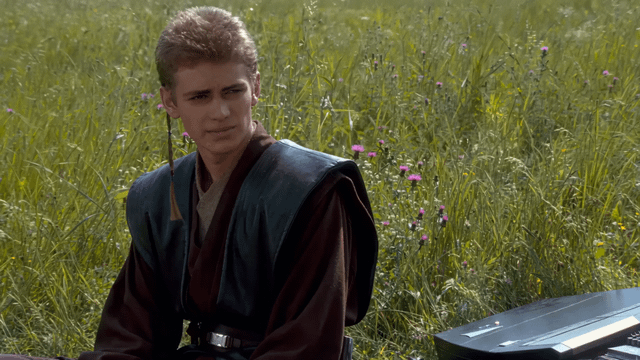 This image features Anakin Skywalker, portrayed by Hayden Christensen, from *Star Wars: Episode II – Attack of the Clones*. He is sitting in a sunlit, grassy meadow dotted with small purple wildflowers, wearing his Jedi Padawan robes complete with a traditional Padawan braid. His expression is pensive, with a furrowed brow and a downturned mouth, conveying quiet inner conflict or contemplation. To his right, a portion of a sleek, futuristic datapad-style device is visible in the frame. This scene is frequently repurposed as a reaction image online to express relatable feelings of doubt, frustration, thoughtful hesitation, or quiet unease about a situation.