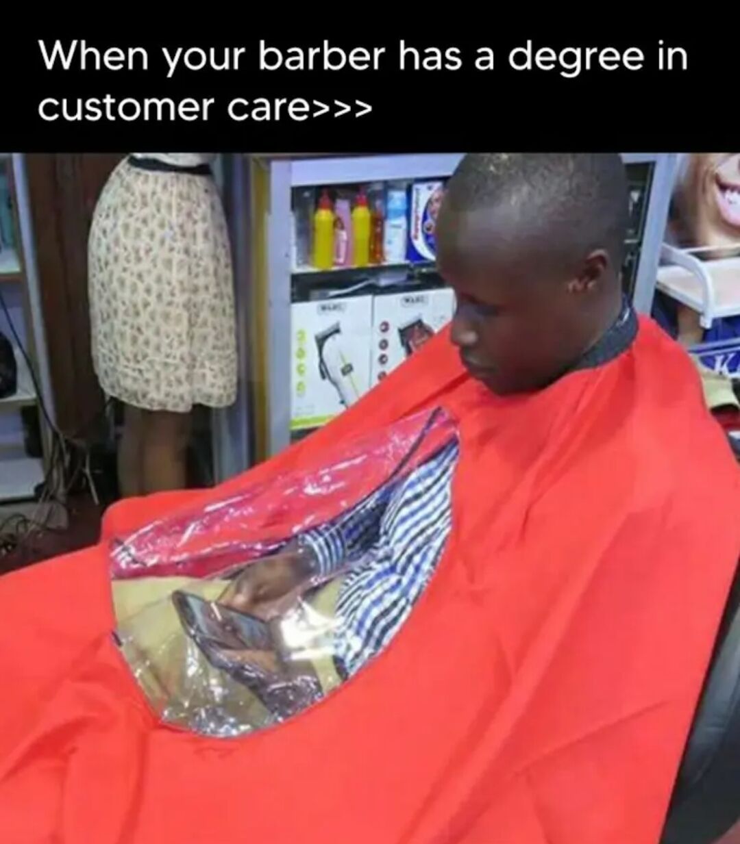 A photograph capturing a person seated in a barber chair, draped in a bright red barber cape. The cape features a clear plastic window over the chest area, revealing the individual's hands—likely holding a phone, as suggested by the shape visible through the plastic. In the background, a shelf stocked with hair care products (yellow bottles, a boxed hair dryer, and other toiletries) is visible, alongside a mannequin dressed in a leopard-print skirt. The top of the image displays the text: 'When your barber has a degree in customer care>>>', humorously implying the barber added the plastic window to allow the customer to use their phone comfortably during the haircut, showcasing thoughtful and attentive service.