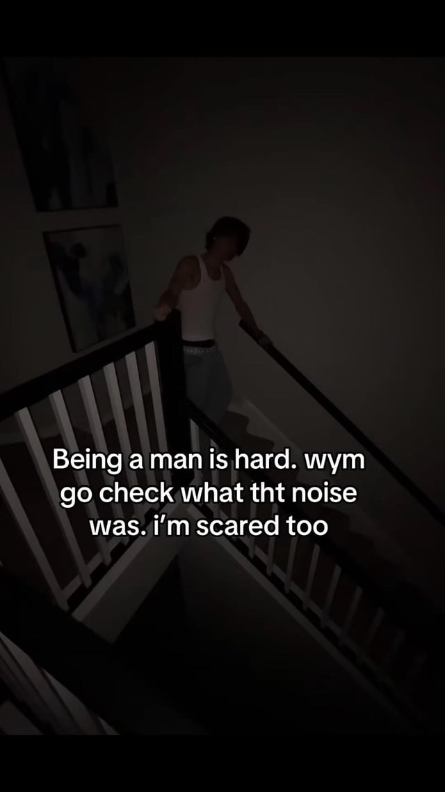 The image shows a dimly lit, dark scene of a young man wearing a white tank top and jeans hesitantly walking down an indoor staircase, holding onto both railings as he descends. White overlaid text on the image jokes about the common gendered social expectation that men are supposed to investigate unknown, potentially scary noises in the home even when they are also just as frightened as everyone else, poking fun at unfair gender roles around being a 'protector'.