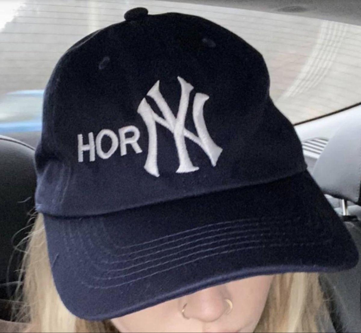 This image features a close-up shot of a person wearing a dark navy bootleg baseball cap modeled after official New York Yankees MLB merchandise. The cap has the iconic interlocking white NY Yankees logo embroidered next to the partial text 'HOR', which combines with the NY logo to unintentionally (or intentionally) form the vulgar word 'HORNY' as a visual pun. Only the lower half of the wearer's face is visible, showing blonde hair and a double nose ring, and the photo is taken from inside a car. The humor comes from the poorly designed counterfeit merch that creates an obscene, unexpected phrase when merging the official brand logo with partial added text.
