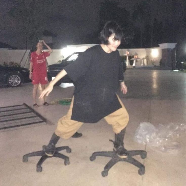 This is a low-light night photograph that creates a humorous optical illusion. A woman wearing an oversized black t-shirt, loose tan shorts, and black boots is balancing with her legs spread wide, each foot placed on a separate rolling, seatless office chair base. From the camera angle, the chair bases look like they are attached directly to her feet, making it appear as if she has giant wheeled feet that allow her to skate or glide across the paved surface. In the background, a second person in a matching red athletic outfit stands near parked cars outside a residential property, holding one hand to their head as they watch the woman.