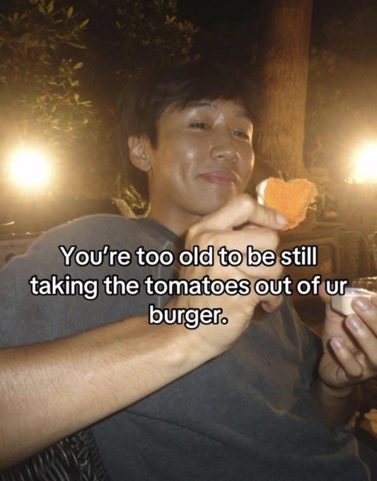 This is a relatable humorous meme featuring a real young East Asian man sitting outdoors at night, illuminated by warm background lights. He has a smug, slightly judgmental amused expression, holding a piece of fried food in one hand and a small sauce container in the other. Overlaid white text on the image reads 'You're too old to be still taking the tomatoes out of ur burger.' The joke targets the common societal expectation that people outgrow picky eating habits like removing disliked ingredients such as tomatoes from their food as they age, teasing people who retain these preferences into adulthood.