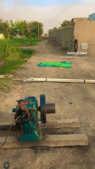 This image shows a dangerous homemade improvised cannon built from a small single-cylinder diesel engine. The cannon is secured to two wooden blocks set on a rural dirt road, and is aimed down the empty road at a green tarp that serves as a target laid out further along the path. White PVC pipes lie on the ground between the cannon and the target, with grass and trees on the left side of the road and low earthen concrete walls on the right. The original Reddit post asks men browsing the subreddit if they would be willing to try this risky homemade experiment.