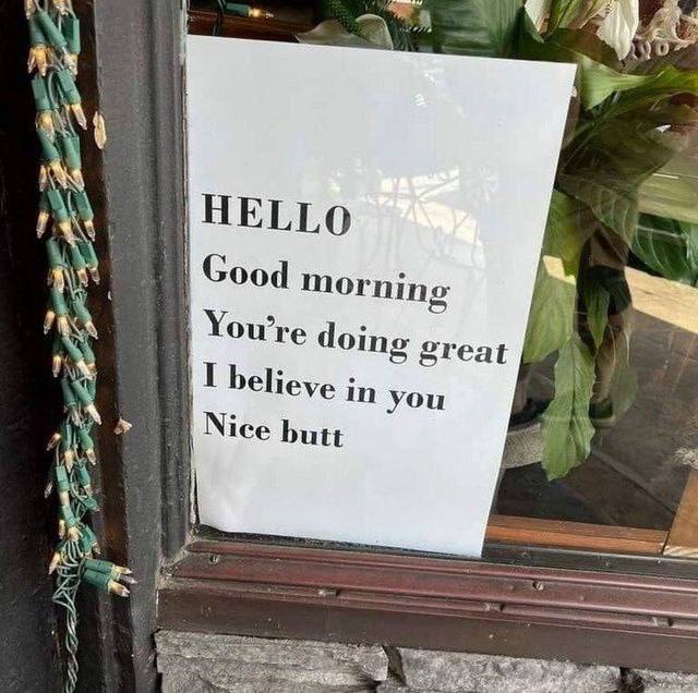 A photograph of a white sign posted in a window, featuring a sequence of friendly, encouraging messages that take a playful turn. The sign has black text reading: 'HELLO', 'Good morning', 'You're doing great', 'I believe in you', followed by the unexpected and humorous 'Nice butt'. The background includes green leafy plants visible through the window, and a string of green and yellow decorative lights hangs on the left side of the frame. The tone shifts from sincere encouragement to lighthearted humor with the final line, creating a funny and wholesome effect.