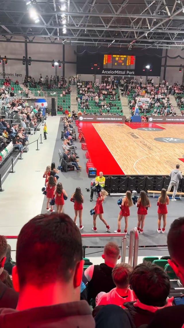 The image captures a live indoor basketball game at the ArcelorMittal Park arena. In the foreground, the backs of audience members' heads are visible, showing they are engaged in watching the event. In the mid-ground, a group of cheerleaders in matching red sequined outfits with blue pom-poms are performing near the edge of the basketball court; one cheerleader is in a crouched, dynamic pose while the others stand in a neat formation. The polished basketball court with red boundary zones is visible, and the arena's tiered seating is filled with spectators. A digital scoreboard hangs above the court displaying game scores, and the 'ArcelorMittal Park' signage is prominently shown on the upper wall. A security staff member in a high-visibility yellow vest stands near the court, and partial text 'Świętochłowice łączy' is visible on the left wall, indicating the arena's location in Świętochłowice, Poland. The scene is lit with event-specific red and white lighting, enhancing the energetic live atmosphere.