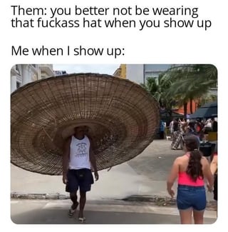 This is a relatable meme using a two-part text setup and a candid photo punchline. The top text first shows a request from others: 'Them: you better not be wearing that fuckass hat when you show up', followed by 'Me when I show up:'. The photo below shows a man walking down a sunny, tropical-looking urban street wearing an absurdly oversized woven straw hat that is several times wider than his entire body, while dressed casually in a white tank top and dark shorts. Other pedestrians are visible in the background. The humor comes from the protagonist intentionally choosing the most extreme, ridiculously oversized version of a hat possible to directly defy the request not to wear a silly hat, leaning into the absurdity far beyond what the requester expected.