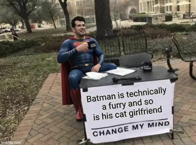 A real person dressed as Superman sits on a small table in a park-like environment. He wears a blue Superman suit with a red cape, red boots, and a yellow belt, and holds a black mug in his right hand. On the table in front of him is a white sign with bold black text that reads: 'Batman is technically a furry and so is his cat girlfriend CHANGE MY MIND'. The background features brick pavement, greenery, benches, and distant buildings, creating a casual outdoor atmosphere. The individual has a slight smile, appearing amused by the sign's content.