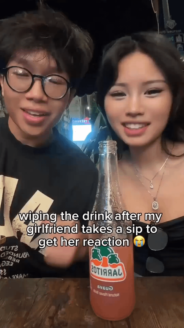 The image features a young East Asian man wearing glasses and a young East Asian woman sitting side by side at a wooden table, both smiling at the camera. A bottle of guava-flavored Jarritos soda sits on the table in the foreground between them. Overlaid white text on the image reads "wiping the drink after my girlfriend takes a sip to get her reaction" next to a crying laughing emoji. The original title of the meme is "I need to find Jesus", a humorous hyperbolic reaction to the petty playful prank shown, where the boyfriend is teasing his girlfriend by pretending he finds her saliva/germs gross enough to wipe the bottle mouth after she drinks from it.