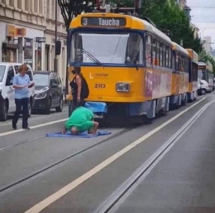 A photograph depicting a city street scene with a yellow tram labeled 'Leipzig' (number 2113, destination 'Taucha') stopped on tracks. In the foreground, a person wearing a green shirt is performing push-ups on a blue mat placed directly on the tram tracks. To the left, two individuals are present: one dressed in black standing near the front of the tram, and another in a light blue shirt standing on the sidewalk holding an object, possibly a phone or notepad. The background includes parked cars, buildings, and trees lining the street, suggesting an urban setting in Leipzig.