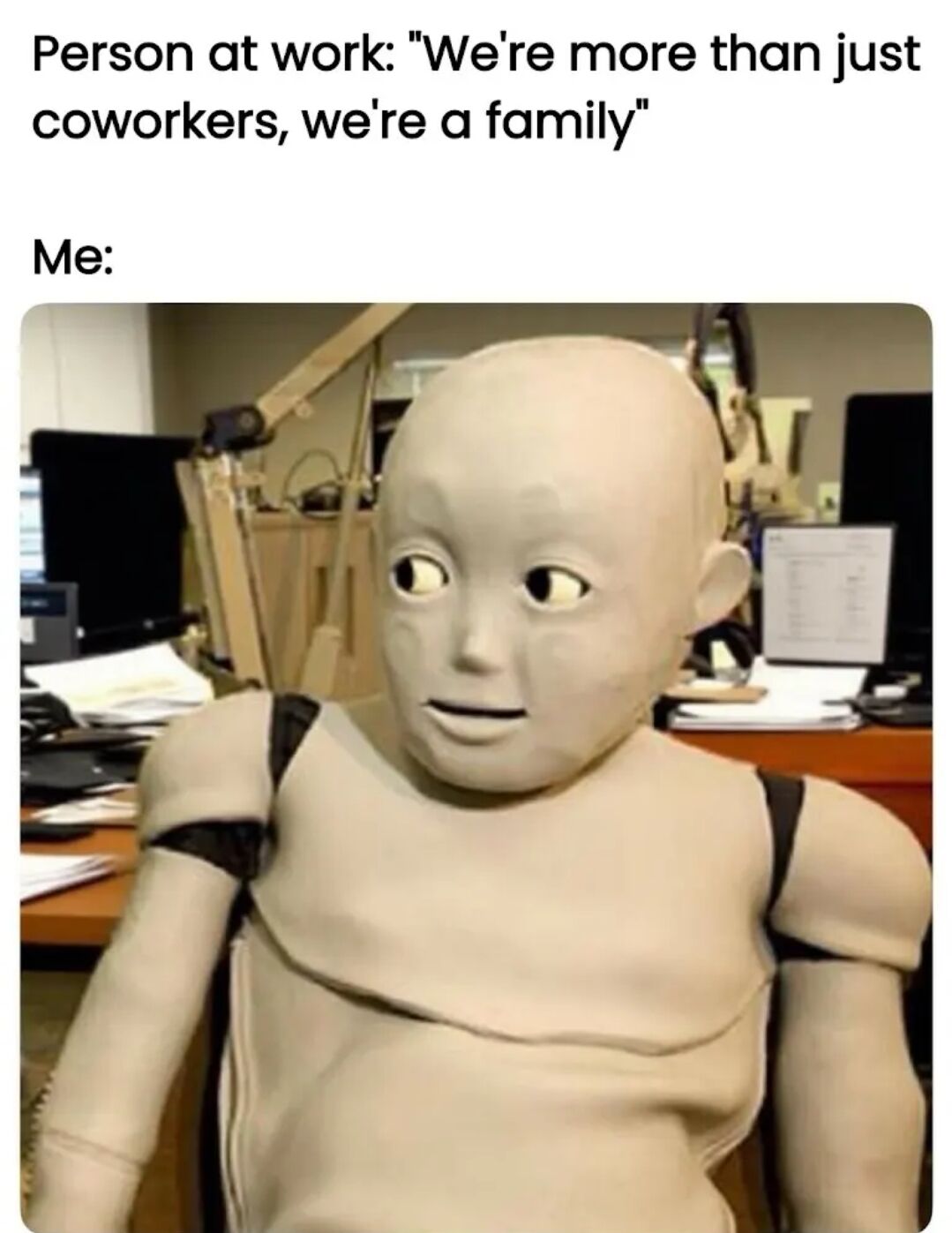 The meme features two text sections followed by an image. The top text reads: 'Person at work: "We're more than just coworkers, we're a family"' and the lower text says 'Me:'. Below this text is a photograph of a humanoid figure with a smooth, pale complexion, large round eyes, and a neutral, slightly unsettling expression, seated in an office environment with computer monitors, papers, and office equipment in the background. The image humorously portrays the reaction to the common workplace cliché of colleagues being referred to as 'family'.