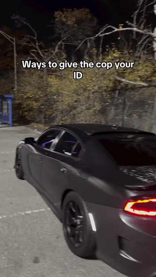 A nighttime photograph of a black car, possibly a sports car, positioned in a dimly lit area with trees and a blue structure visible in the background. The image has a text overlay at the top that reads 'Ways to give the cop your ID', suggesting a humorous or sarcastic context related to interactions with law enforcement during traffic stops where identification is requested.