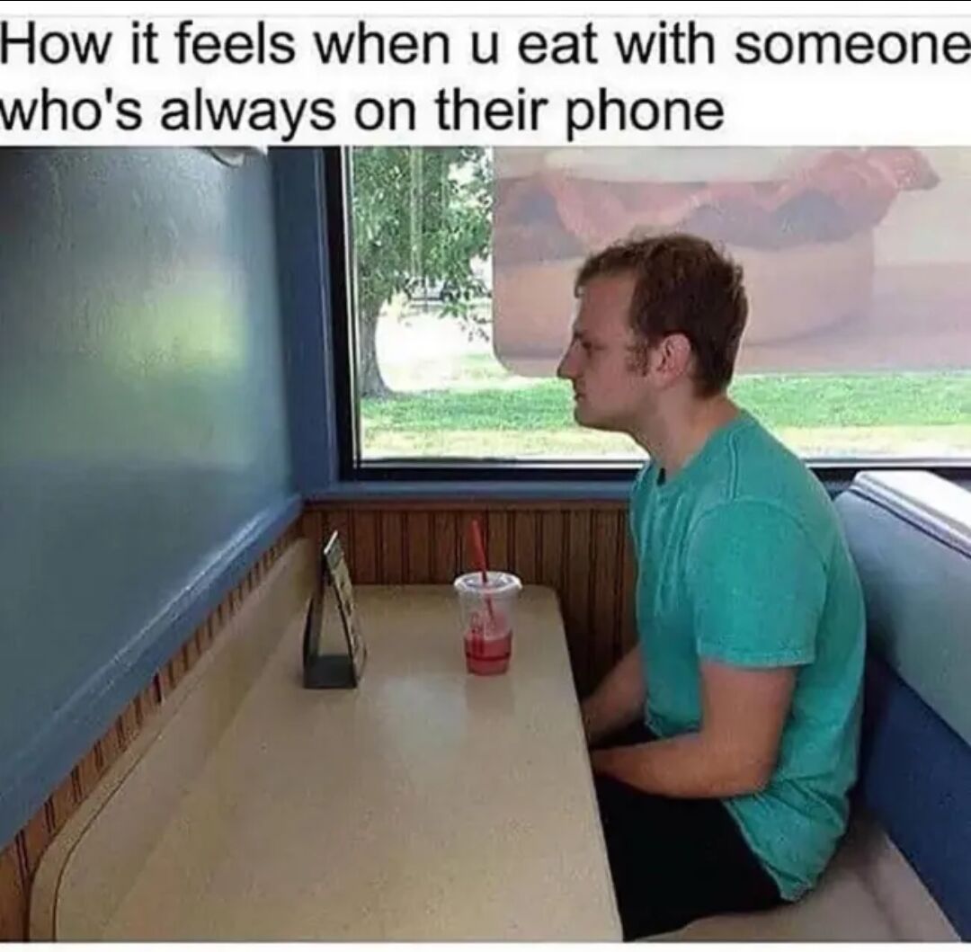 This meme shows a young man in a turquoise short-sleeved t-shirt sitting alone on one side of a diner booth. He leans slightly forward, looking bored and awkward as he stares at the empty seat across the table, which only holds a single red slushie in a plastic cup and a small table stand. The text at the top of the image reads 'How it feels when u eat with someone who's always on their phone', humorously capturing the relatable feeling of loneliness and awkwardness when a dining companion ignores you completely to use their phone, making you feel as if you are eating alone even when they are physically present.
