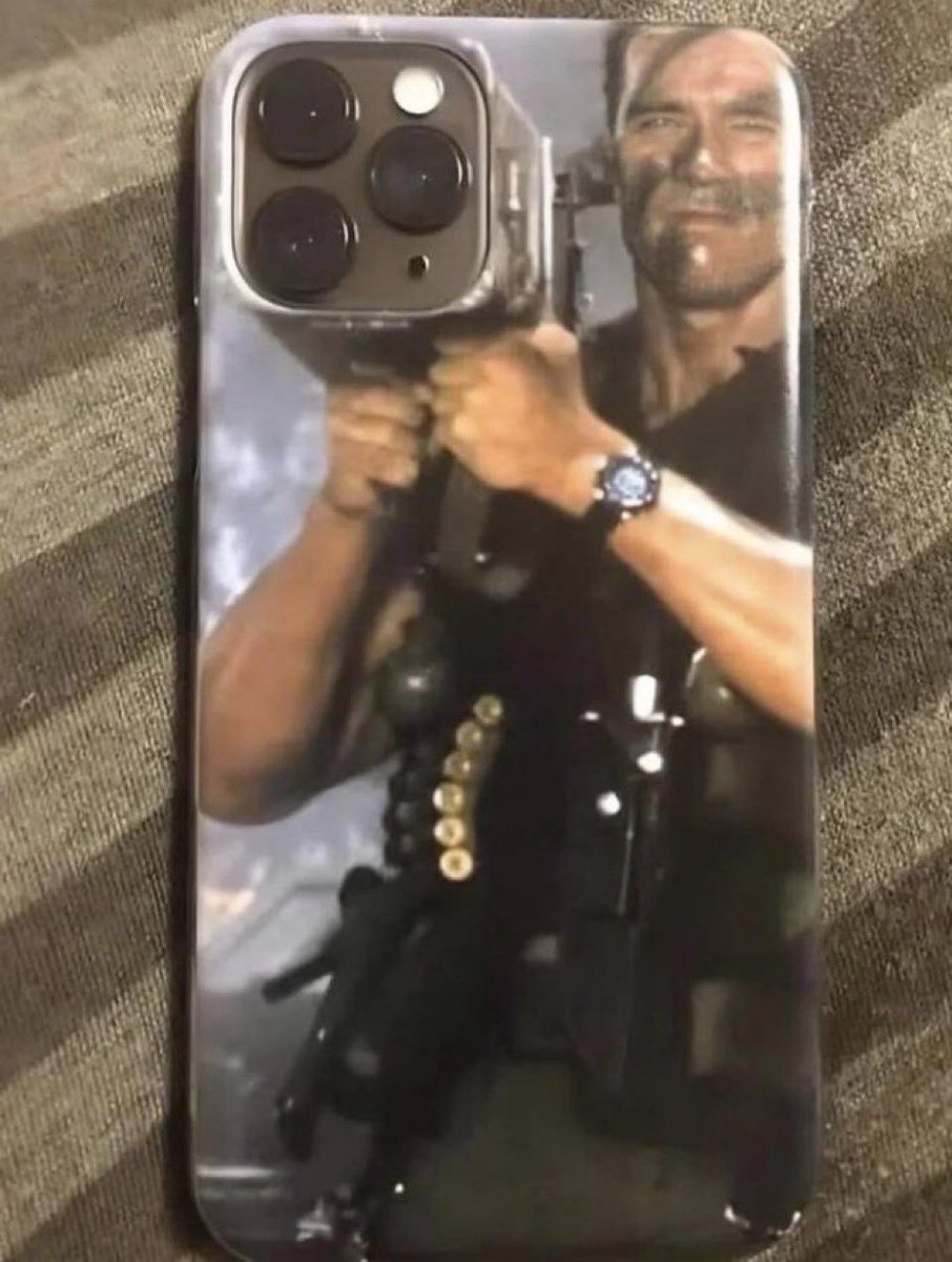 This image shows a custom printed iPhone case featuring a still of Arnold Schwarzenegger as John Matrix from the 1985 action movie Commando, holding a large firearm aimed directly forward. The design is intentionally positioned so the iPhone's rear triple camera bump lines up perfectly with the barrel of the gun Schwarzenegger is holding, creating a humorous visual effect that makes it appear as if the phone's camera lenses are the end of the weapon barrel he is aiming.